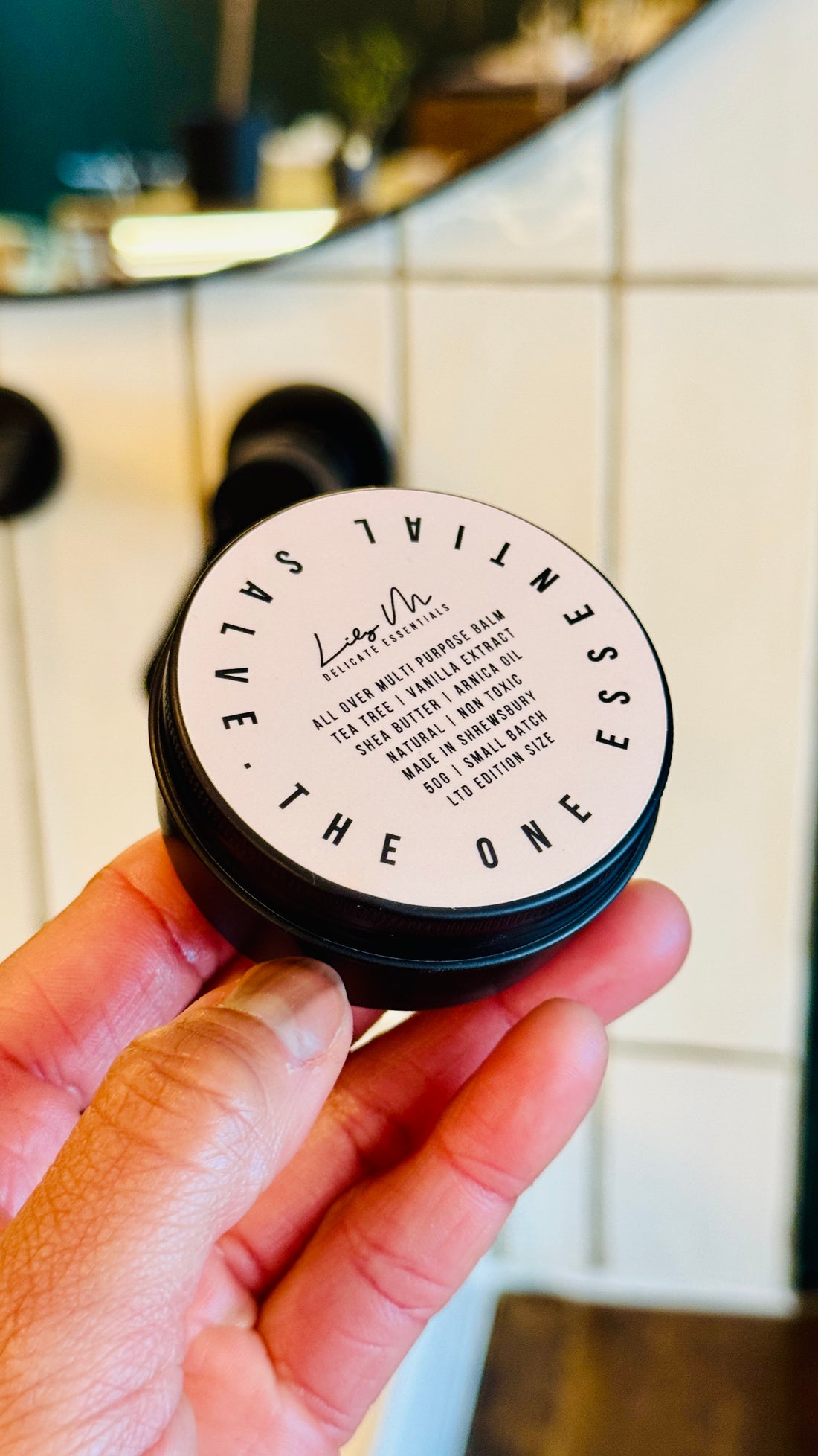 Multi purpose Balm being held, with the lid on showing the front label, which says - All over multi purpose balm tea tree vanilla extract shea butter arnica oil natural non toxic made in Shrewsbury 50g small batch Ltd edition size