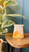 wax melt burner with a wax melt in the top burner away with a candle lit underneath, a plant to the left hand side on the image.