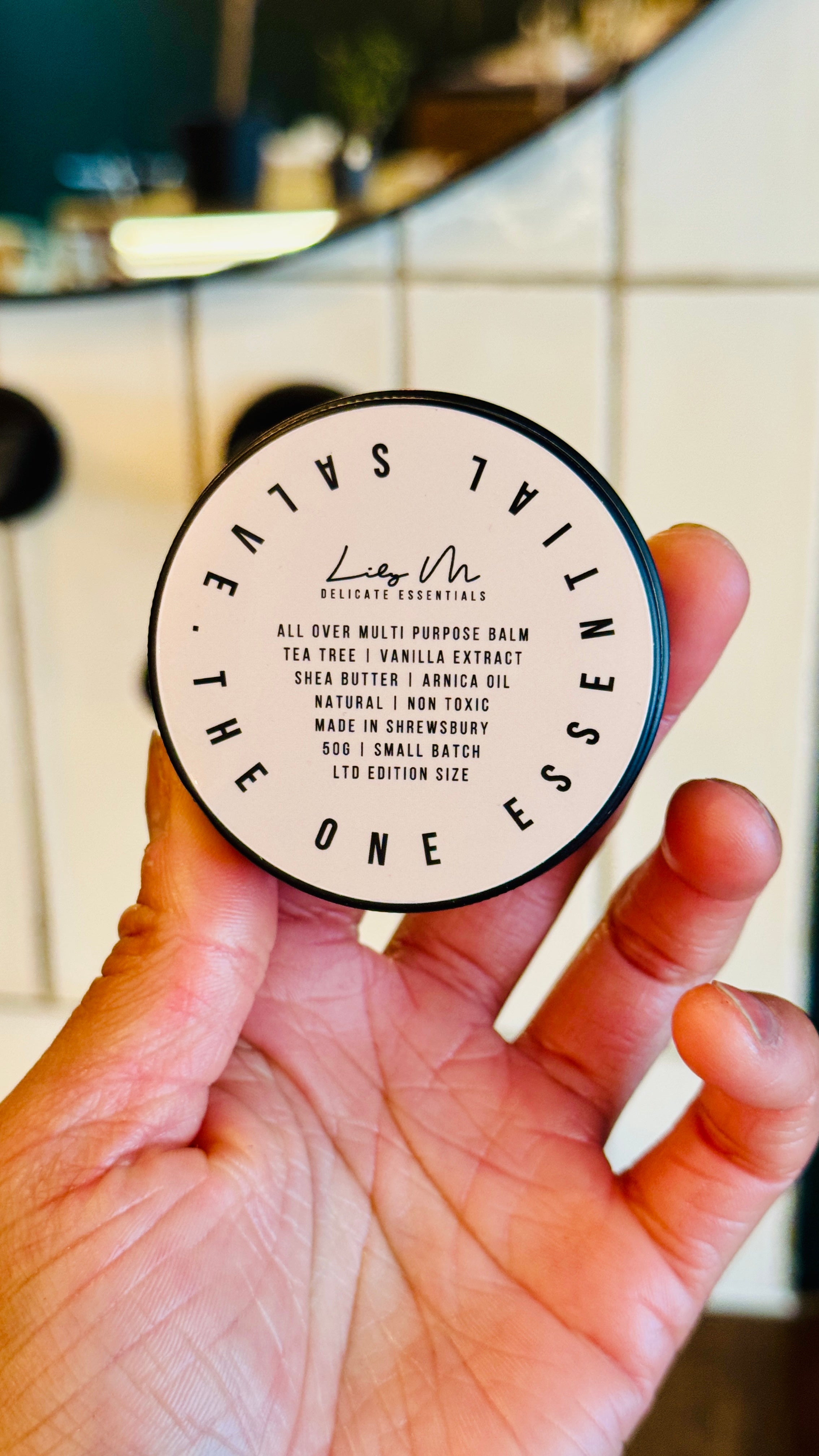 Multi purpose Balm being held, with the lid on showing the front label, which says - All over multi-purpose balm tea tree vanilla extract shea butter arnica oil natural non toxic made in Shrewsbury 50g small batch Ltd edition size