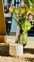 handmade white glaze candle jar on a hessian rug on the window cill with a vase of flowers behind