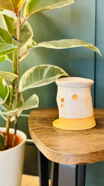 handmade wax melt burner in a white glaze, turn to the left on a wooden table with a plant in the left side of the image