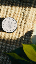 multi purpose balm with lid on, placed on a hessian rug, plant in the bottom right hand corner