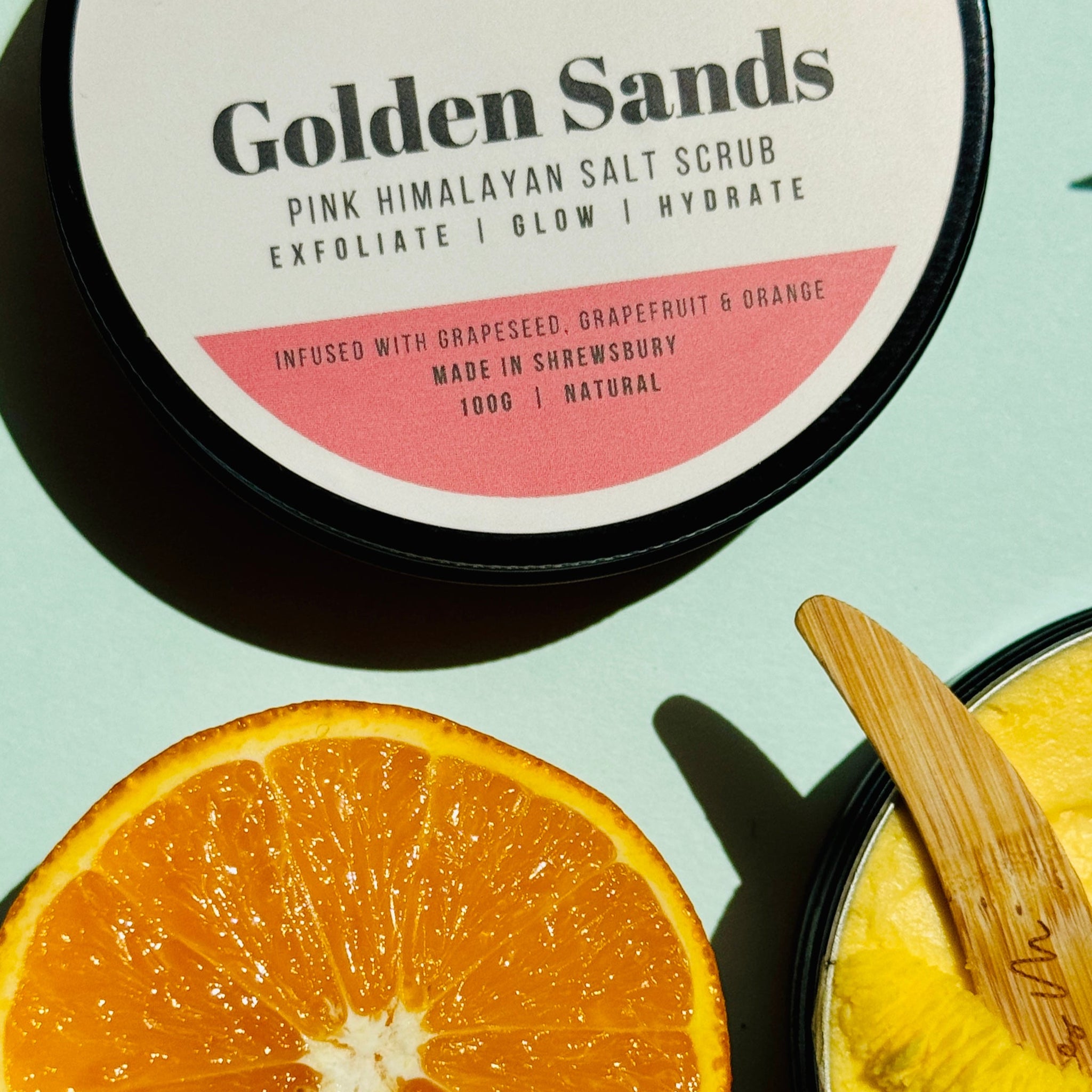 Orange sliced with open tub of bay butter and a scoop and a closed tin of salt scrub - golden sands 