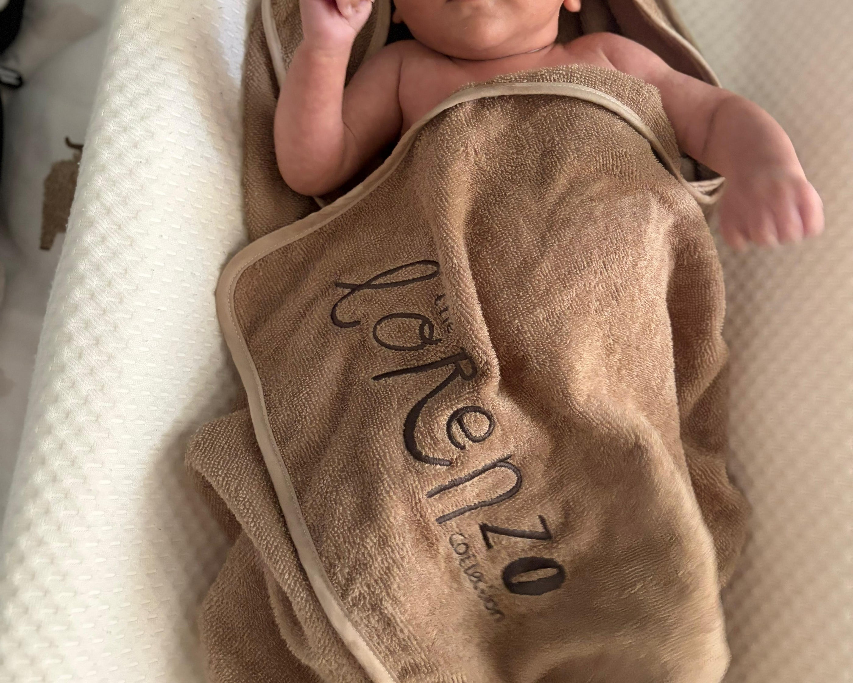 A newborn baby wrapped in a tan hooded towel with bear ears and the brand name 'lorenzo' printed on it, lying on a textured white bassinet.
