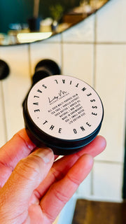 Multi purpose Balm being held, with the lid on showing the front label, which says - All over multi purpose balm tea tree vanilla extract shea butter arnica oil natural non toxic made in Shrewsbury 50g small batch Ltd edition size 