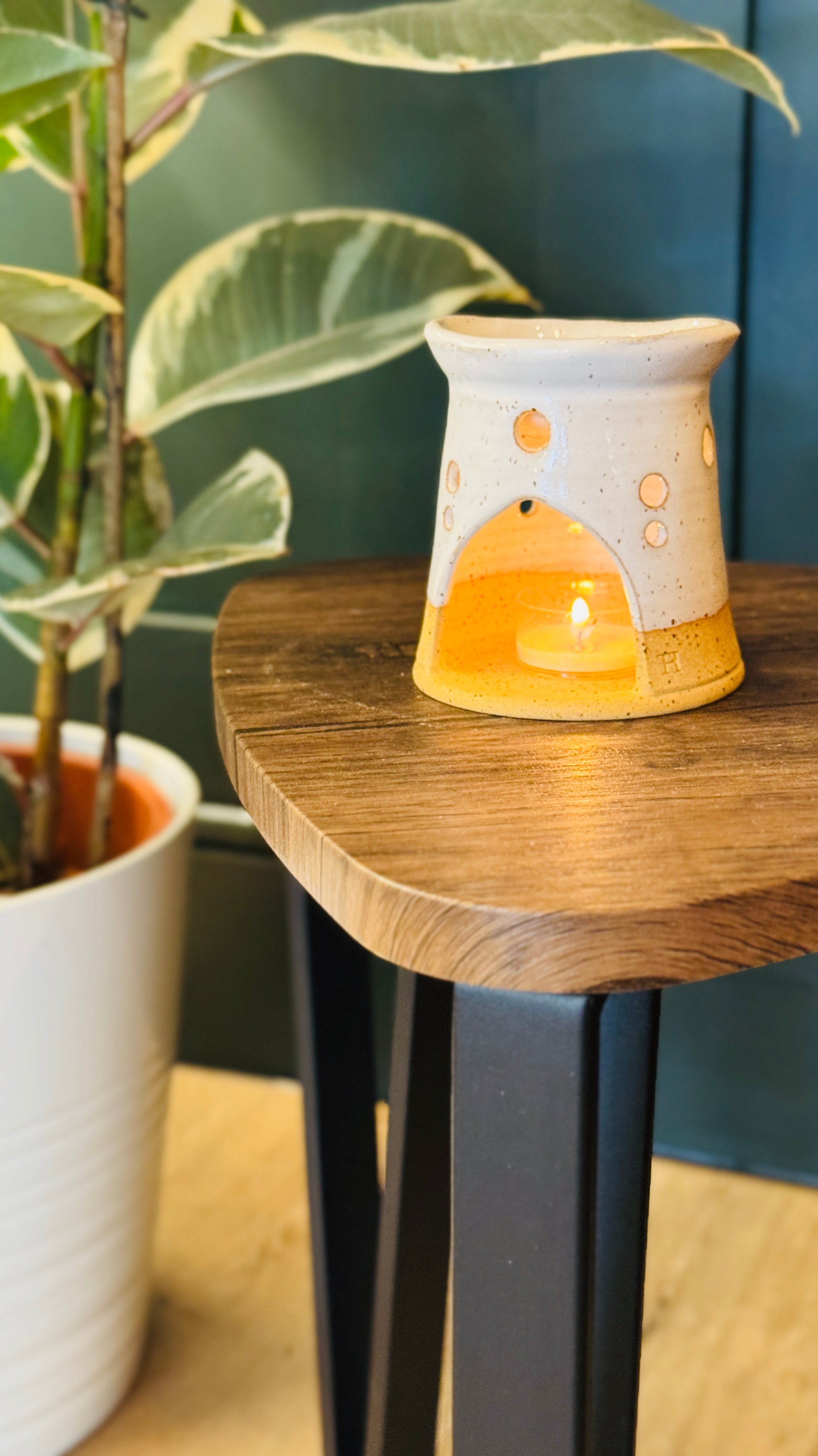 handmade wax melt burner in a white glaze, facing the front with a candle lit, on a wooden table with a plant in the left side of the image 