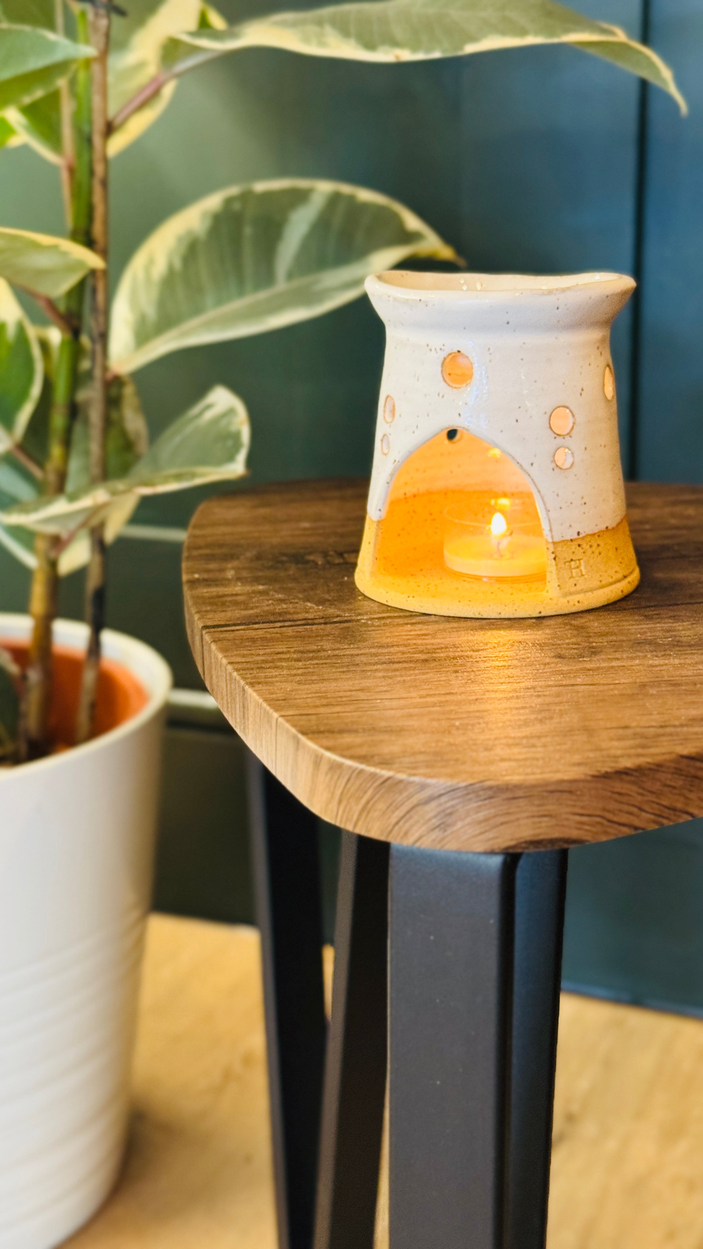 handmade wax melt burner in a white glaze, facing the front with a candle lit, on a wooden table with a plant in the left side of the image 