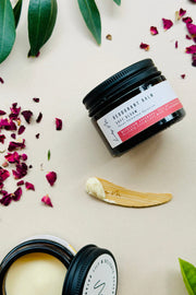 Two black jars of deodorant cream with a wooden applicator on a light background with green leaves and pink petals.