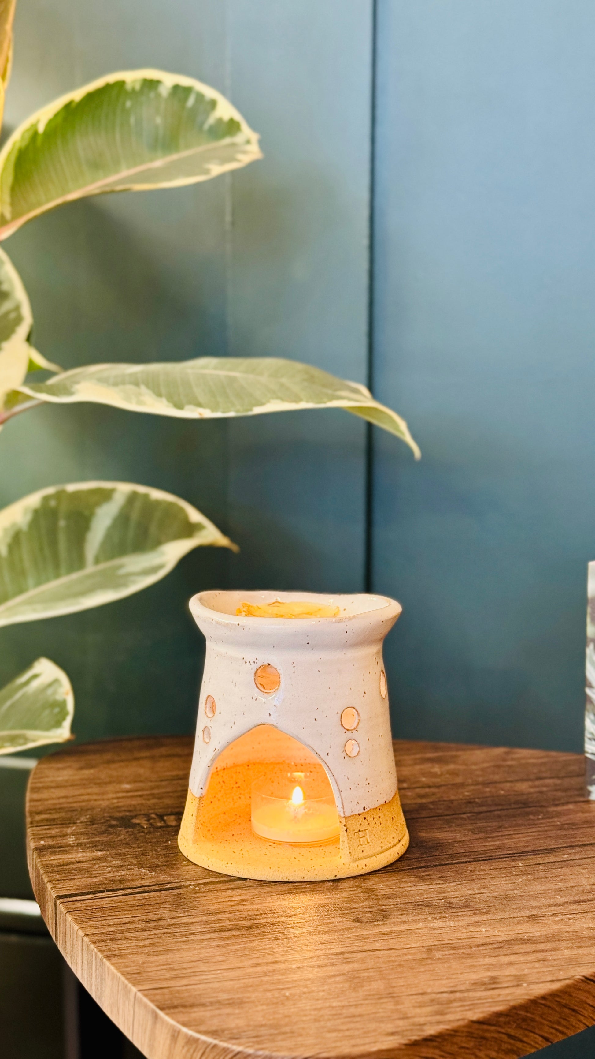 handmade wax melt burner in a white glaze, facing to the front with a wax melt burning  on a wooden table with a plant in the left side of the image 
